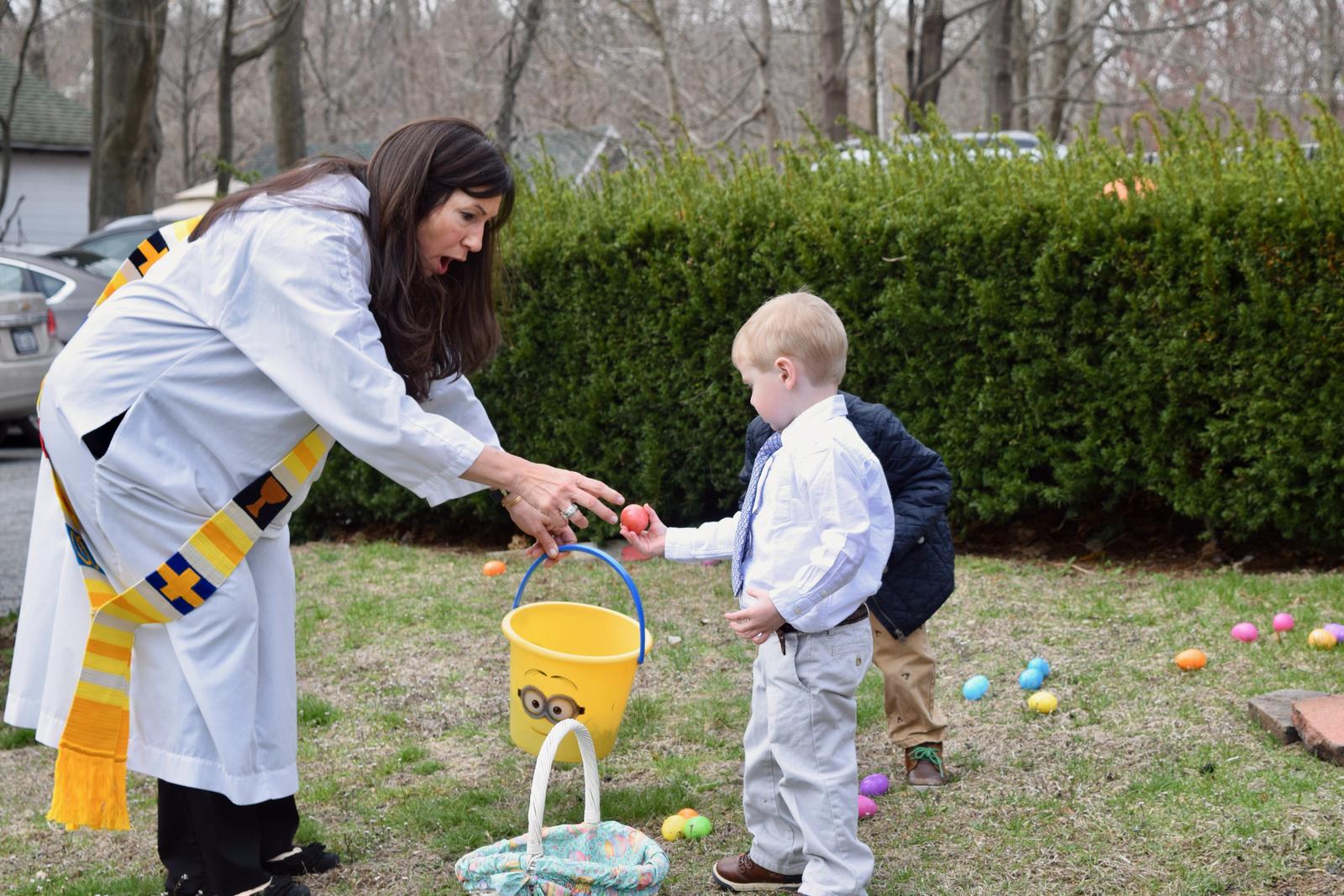 A woman in a white robe and colorful stole hands a little boy an Easter egg during the egg hunt
